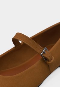 Παντόφλα Mary Jane από καφετί σουέντ με ραμμένο λουράκι και μικρή ασημί πόρπη κλεισίματος σε λευκό φόντο.