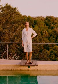 Femme en blazer blanc et short jusqu'aux genoux, pieds nus au bord de la piscine, tenant un petit sac, avec en arrière-plan des arbres verts denses.