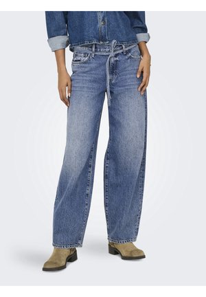 Frau trägt locker sitzende ausgewaschene blaue Jeans mit einem Stoffgürtel, der in der Taille gebunden ist, beige Stiefeletten mit Absatz und eine hochgekrempelte Jeansjacke.
