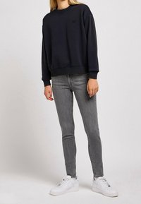 Felpa blu marino con scollo rotondo e polsini a costine, abbinata a jeans aderenti grigi e scarpe da ginnastica bianche. Presenta un logo sul petto.