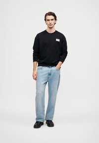 Felpa nera con logo, jeans dritti azzurri chiari e scarpe nere. L'outfit presenta una vestibilità comoda e un design semplice.