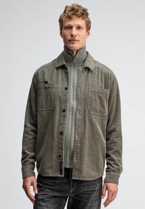 Olive green corduroy shirt jacket with two front pockets, button closure, and ribbed texture, layered over a light green zip-up top.