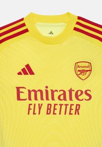 Žltý futbalový dres Arsenalu s červenými pruhmi a logami Adidas, erbom klubu Arsenal a reklamou "Emirates Fly Better".