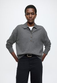 Pulover gri tricotat cu guler, cu o pată de nasturi și manșete cu dungi, asortat cu pantaloni închiși la culoare și curea.