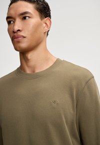 Oliwkowy bawełniany sweatshirt z okrągłym dekoltem, ściągaczami na rękawach oraz haftowanym kwiatowym logo po lewej stronie klatki piersiowej. Gładka tekstura.