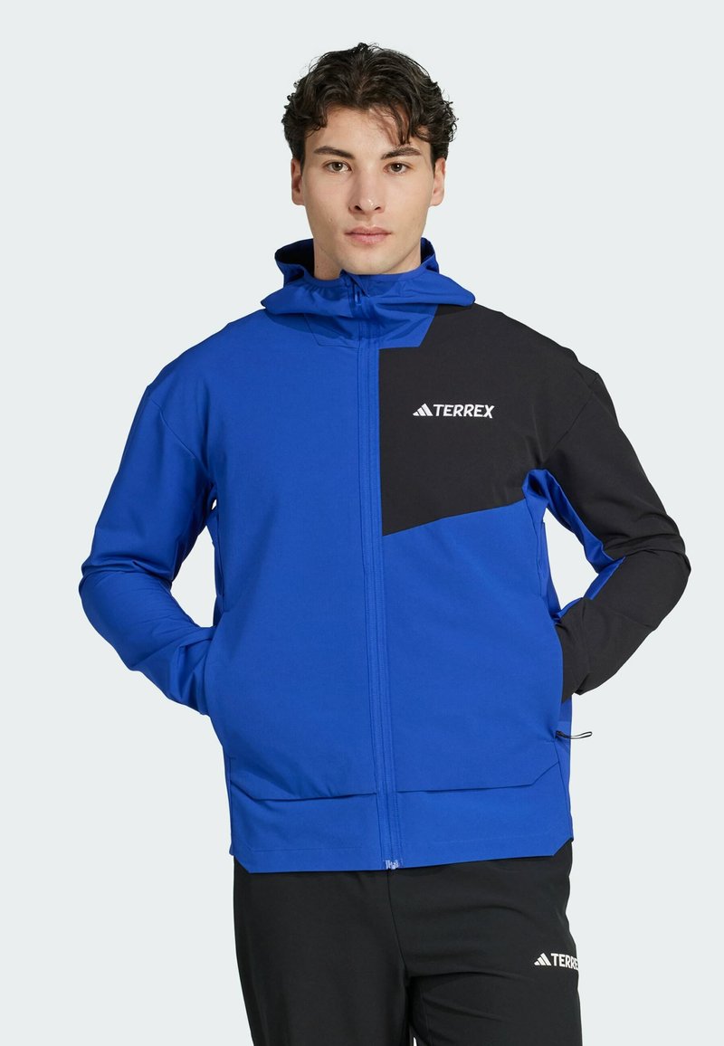 Blauwe en zwarte ritsjack met capuchon, met een groot zwart paneel en wit logo op de borst. Soepele stof, functioneel ontwerp.