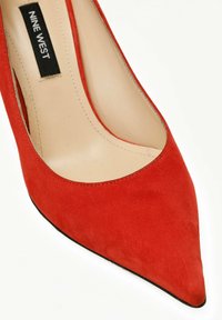 Zapatos de tacón en ante rojo con punta afilada, un escote bajo y suela negra. Presentan una superficie suave y texturizada, así como una etiqueta interna de marca.