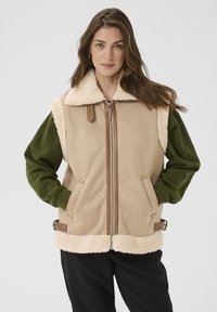 Beige mouwloos vest met een zachte, schapenvacht kraag en voering, groene mouwen en bruine hardware. Beschikt over zijzakken en verstelbare bandjes.
