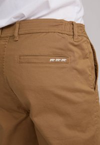 Pantalons en coton marron avec une poche arrière ornée d'un motif embossé de trois vaches. Texture lisse et coupe ajustée.