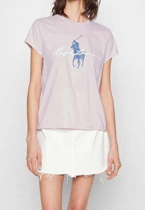 Jasnofioletowa bawełniana koszulka z krótkim rękawem, z graficznym motywem niebieskiego gracza polo i białym napisem, w połączeniu z białą spódnicą dżinsową.