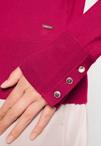 Fuchsia gebreide trui met geribbelde manchetten en drie metalen knopen. Kleine metalen logoplaat op de borst. Gladde textuur, nauwsluitend ontwerp.