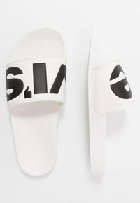 Sandalias deslizantes blancas con una ancha tira de logo negro. El material suave combina una textura lisa con un diseño minimalista. Suela plana.