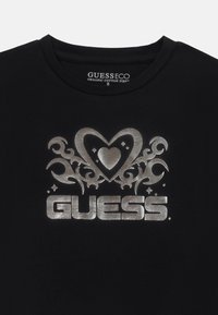 Svart bomull t-shirt med en silvergrafik som föreställer ett hjärta och dekorativa svängar, tillsammans med "GUESS" logotypen i fetstil.