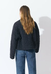Veste noire matelassée au design court, avec un motif géométrique et des poignets côtelés, assortie à un jean en denim bleu clair.