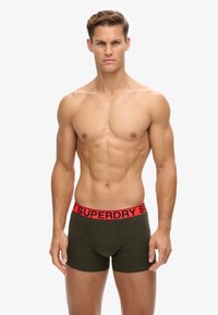 Grüne Boxer-Shorts mit einem roten Bund, der die "SUPERDRY"-Markierung trägt. Der Stoff wirkt weich, hat eine enganliegende Passform und keine sichtbaren Muster.