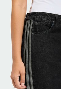 Čierne džínsy z denimu s tromi zvislými sivými pruhmi na boku, s priliehavým pásom a prednými vreckami. Na páse je malý detail s logom.