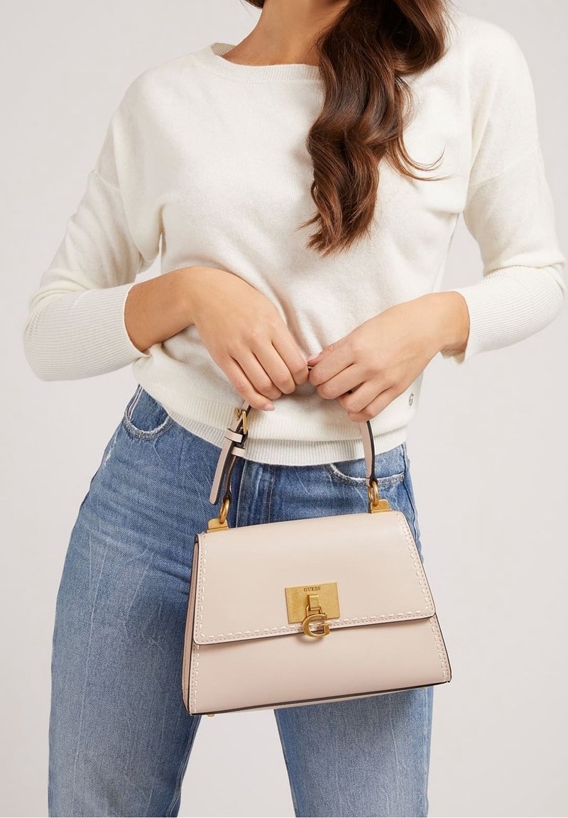 Femme portant un pull blanc et un jean bleu tenant un sac à main en cuir beige avec des éléments dorés et une poignée supérieure devant un fond uni.