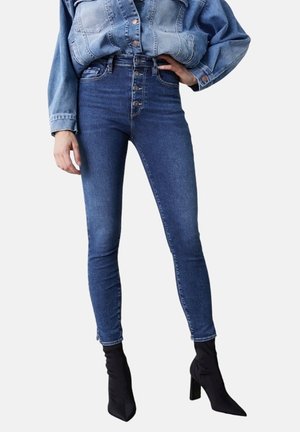 Hochwaisted blaue Denim-Jeans mit einer Fünf-Knopf-Front, taillierter Passform und minimalen Abnutzungen, kombiniert mit schwarzen Stiletto-Stiefeletten.