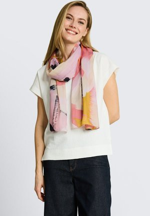 Uśmiechnięta kobieta w białym topie z krótkim rękawem, ciemnych dżinsach i wielobarwnym pastelowym szaliku na jasnym tle.
