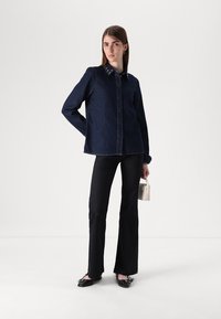 Camicia in denim scuro con colletto button-down, adornata con borchie decorative, abbinata a pantaloni neri aderenti e scarpe flat. Borsa bianca minimalista.