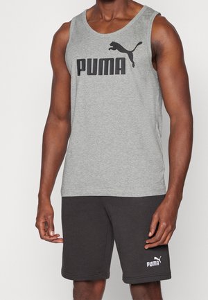 Mężczyzna ubrany w szary top Puma i czarne spodenki Puma, stojący z rozluźnionymi ramionami wzdłuż ciała na jednolitym tle.