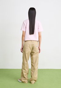 Ljusrosa kortärmad t-shirt i kombination med tanfärgade löst sittande byxor med fickor och en loggopatch. Långt, rakt svart hår synligt.