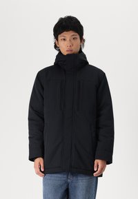 Black puffer jacket with a high collar, front zip, and dual chest pockets. Features a matte finish and casual fit. Worn with blue jeans.