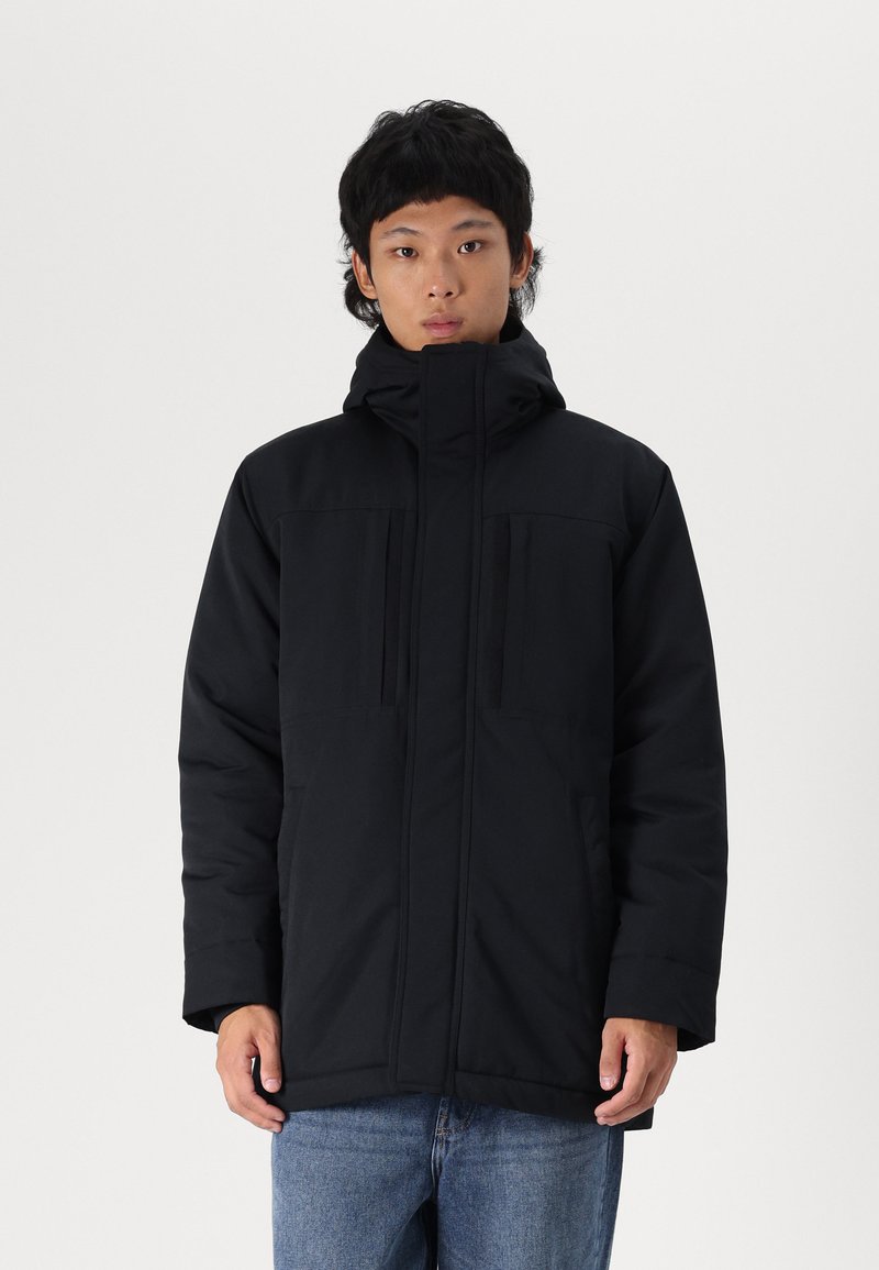 Black puffer jacket with a high collar, front zip, and dual chest pockets. Features a matte finish and casual fit. Worn with blue jeans.