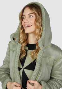 Junge Frau mit welligem, blonden Haar, die eine grüne Lammfell-Häkeljacke und einen schwarz-grünen geometrischen Pullover trägt, lächelt und schaut nach links.