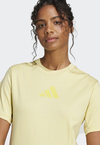 Žluté bavlněné tričko s krátkým rukávem a texturovaným logem značky ve slabě tmavší žluté barvě na přední straně.