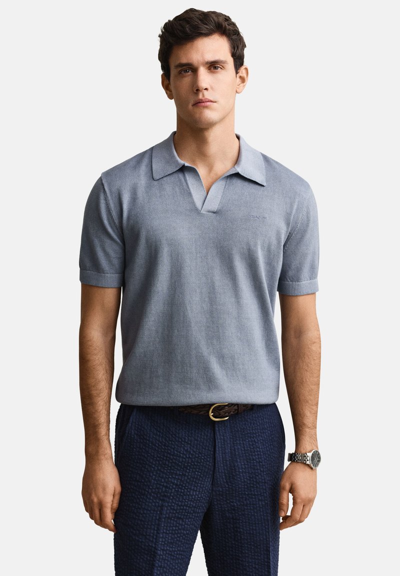 Hombre joven con una camiseta polo de manga corta azul claro, pantalones azul marino texturizados, un cinturón marrón y un reloj de pulsera plateado, de pie en posición neutral.