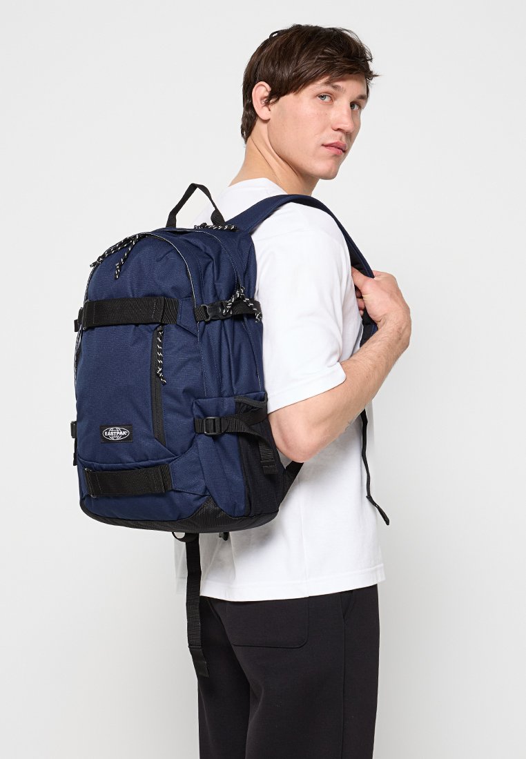 Jeune homme portant un t-shirt blanc et un pantalon noir porte un grand sac à dos bleu marine sur une épaule devant un fond uni.