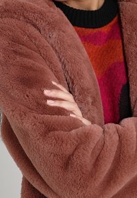 En person som bär en mjuk, brun fuskpälsjacka över en färgglad röd och svart mönstrad tröja, med armarna i kors och välvårdade naglar.