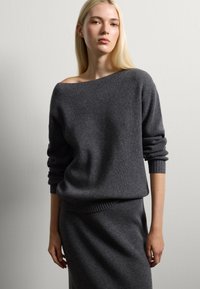 Giovane donna con lunghi capelli biondi che indossa un maglione a coste grigio scuro con spalle scoperte e una gonna coordinata su uno sfondo bianco uniforme.