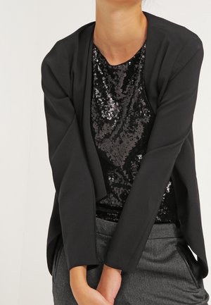 Personne portant un haut noir à sequins sous un blazer noir ouvert sur le devant et un pantalon gris, les bras croisés devant.