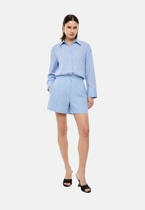 Donna in piedi che indossa una camicia azzurra con bottoni e pantaloncini coordinati con sabot neri col tacco, mani nelle tasche, sfondo neutro.