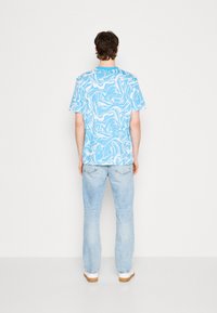 Young man standing facing away, wearing a blue and white abstract patterned short sleeve shirt, light blue jeans, and white shoes.