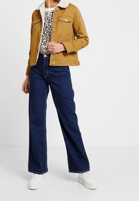 Żółta musztardowa kurtka dżinsowa z białym kołnierzem z baranka, w połączeniu z topem w panterkę i szerokimi, ciemnoniebieskimi dżinsami, do tego białe trampki.