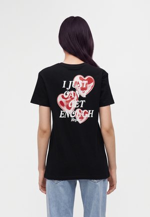 Donna con capelli lunghi e scuri che indossa una t-shirt nera con cuori rossi e testo "I Just Can't Get Enough" sulla schiena, abbinata a jeans azzurri.