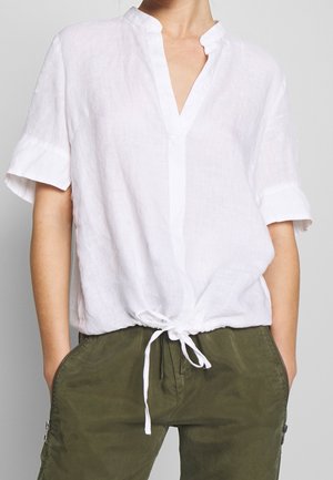 Blusa de lino blanca de manga corta con escote en V y detalle de lazo en la parte delantera, con puños enrollados y una textura ligera.