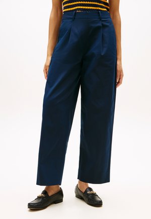 Pantaloni navy a gamba larga indossati con mocassini neri con dettagli a catena dorata e un top senza maniche a righe nere e gialle.