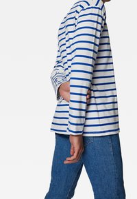 Vit långärmad skjorta med blå horisontella ränder, rundad halsringning, avslappnad passform och sidslitsar vid fållen, tillsammans med jeans.