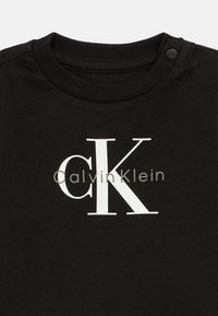 Čierne tričko s veľkými bielymi písmenami "cK" a textom "Calvin Klein" na prednej strane, s gombíkovým zapínaním pri výstrihu.
