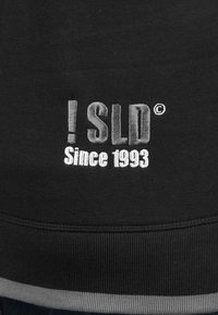 Felpa nera con testo ricamato "! SLD" e "Dal 1993". Testo in fili bianchi e argentati su una superficie di tessuto liscia.