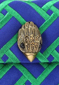 Tessuto blu con cuciture geometriche verdi e un fermaglio decorativo a forma di testa di uccello dorata con occhi neri e dettagli borchiati.