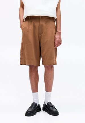 Pantaloni scurți din in maro, cu o croială lejeră, curea laterală și închidere cu nasture. Purtați cu mocasini din piele neagră și șosete albe.