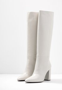 Botas de cuero blancas hasta la rodilla con una textura suave, punta afilada y un tacón grueso alto. Los detalles de las costuras añaden un sutil interés de diseño.