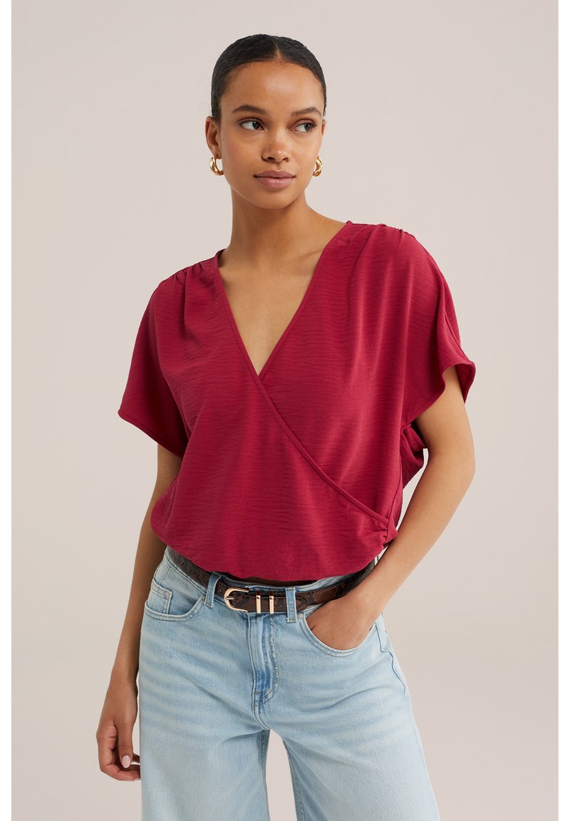 Femme aux cheveux foncés en chignon bas portant des boucles d’oreilles créoles dorées, un chemisier cache-cœur rouge à col en V, un jean bleu clair et une ceinture marron, avec une main dans la poche.