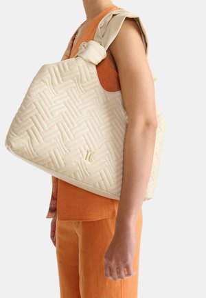 Donna che indossa un top arancione senza maniche e pantaloni porta una grande borsa trapuntata color crema con manici annodati sulla spalla.