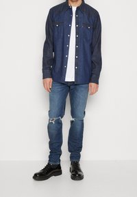 Camisa de mezclilla azul oscuro con dos bolsillos en el pecho, botones a presión y un yugo de estilo western. Combinada con jeans azules desgastados y botas negras.